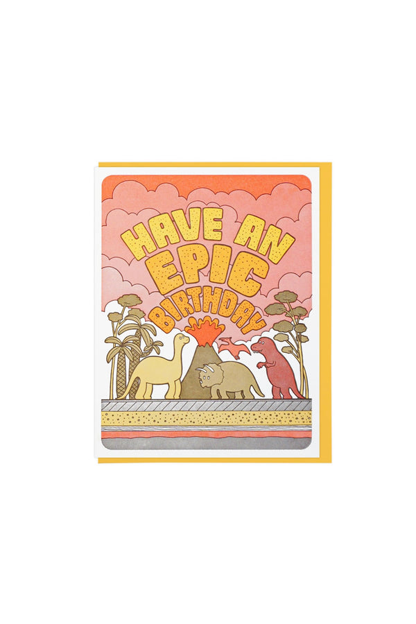 White greeting card with yellow envelope on a white background. The card features three dinosaurs standing in front of an erupting volcano and palm trees beneath an orange cloudy sky. The card says "Have an Epic Birthday" in yellow orange block letters.