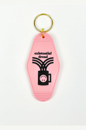 A pale pink motel-style keychain on a white background. The keychain has a gold ring at the top and is decorated with a black image of a coffee cup with an upside down smiley face. The text reads "Existential dread."