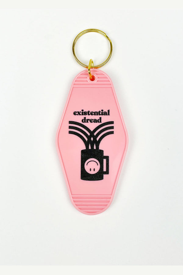 A pale pink motel-style keychain on a white background. The keychain has a gold ring at the top and is decorated with a black image of a coffee cup with an upside down smiley face. The text reads "Existential dread."