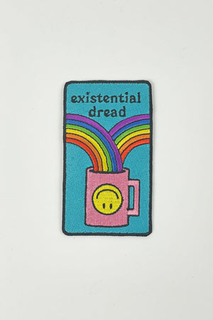 A multi-colored iron-on patch is on a white background. The patch features a black outline around a teal background. A pink coffee cup decorated with a yellow upside down smiley face has two rainbows flowing into it.  The text reads "Existential dread."