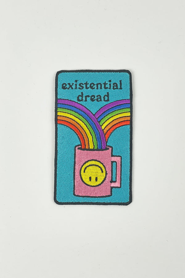 A multi-colored iron-on patch is on a white background. The patch features a black outline around a teal background. A pink coffee cup decorated with a yellow upside down smiley face has two rainbows flowing into it.  The text reads "Existential dread."