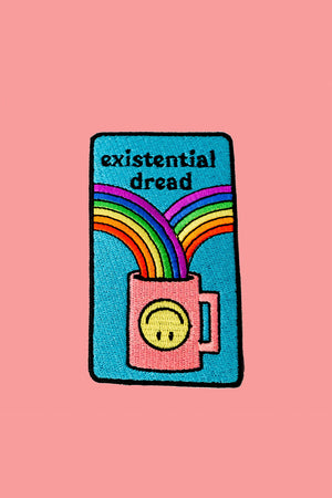 A multi-colored iron-on patch is on a peach colored background. The patch features a black outline around a teal background. A pink coffee cup decorated with a yellow upside down smiley face has two rainbows flowing into it. The text reads "Existential dread."