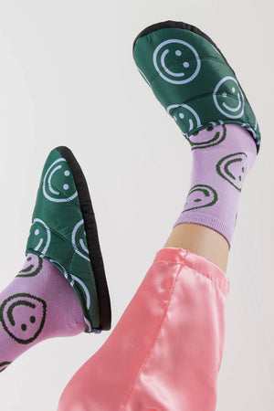A person with purple socks and pink pants  wearing green puffy forest green house slippers with a black rubber sole. The slippers have a light blue smiley face pattern.