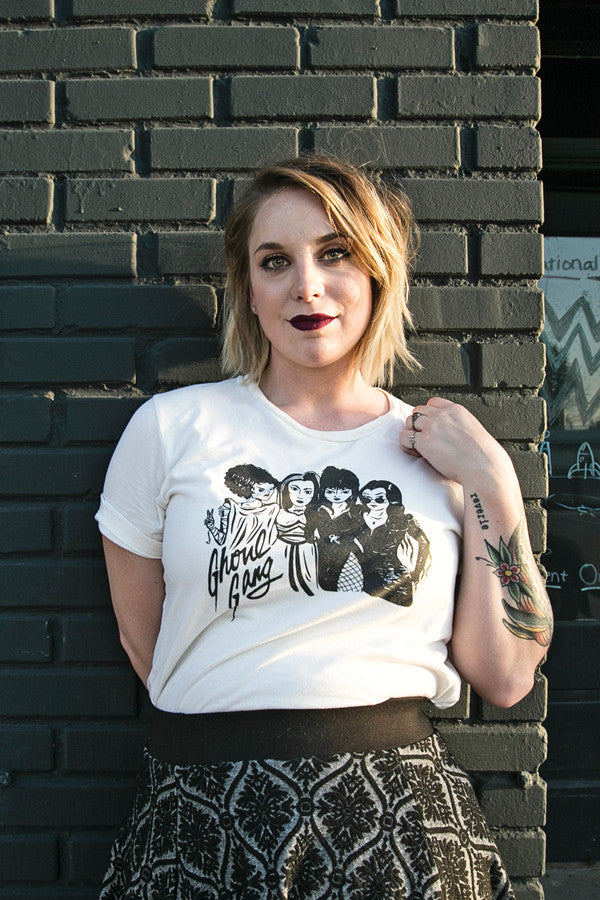 A light-skinned woman with short blonde hair is wearing a Ghoul Gang t-shirt up against a black brick wall.