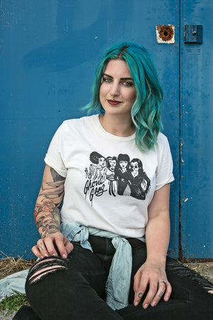 A light-skinned woman with medium-length turquoise hair is wearing an off-white t-shirt with the words 