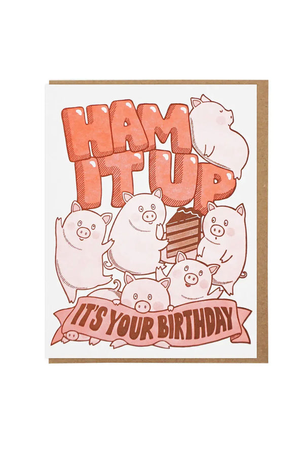White card with kraft brown envelope on a white background. The card features six pink pigs having a piece of chocolate cake surrounding the words "Ham It Up" in light orange bubble letters.  Below the bubble letters and pigs is one pig holding a pink banner that says "It's Your Birthday".