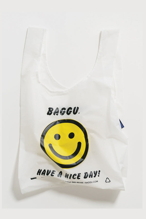 White reusable grocery bag on a white background. The bag features a yellow smiley face with "Baggu" above the face and below it says "Have A Nice Day!" in black lettering.