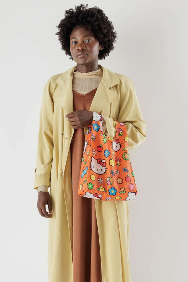Person in front of a white background holding a small orange tote bag featuring Hellow Kitty, Mulit color daisy flowers, red mushrooms, and apples printed all over it. 