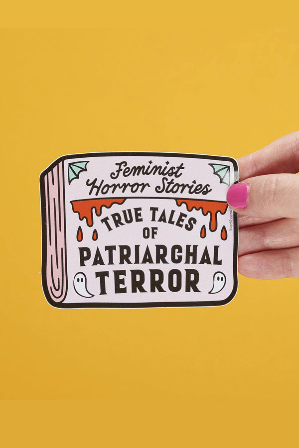 A hand is holding up a rectangular sticker that looks like the cover of a newspaper. It reads "Feminist Horror Stories: True Tales of Patriarchal Terror" with images of ghosts, cobwebs, and blood. The photo background is yellow.