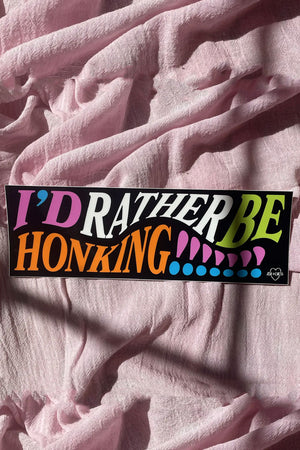 A black bumper sticker with colorful text that reads "I'd rather be honking!!!!!!!" laid on a pink linen backdrop.
