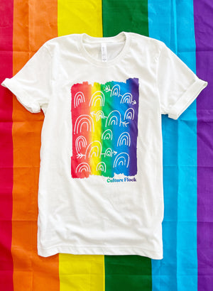 A white t-shirt with a rainbow gradient, decorated with illustrated rainbows in the center is laying on top of a rainbow flag background. The shirt has the words "Culture Flock" near the bottom right corner of the design.