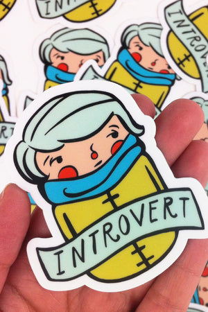 A hand holds a die-cut sticker of an illustrated whimsical person in a mustard yellow overcoat and teal scarf with rosy cheeks. Across their chest is a banner that reads "Introvert." In the background is a collage of identical stickers on a white flat surface.