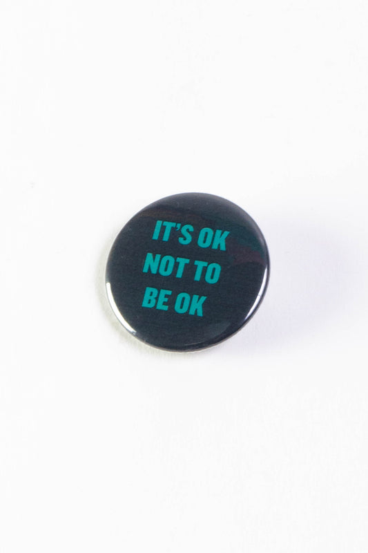 A black pinback button with text that reads "It's ok not to be ok" in teal.