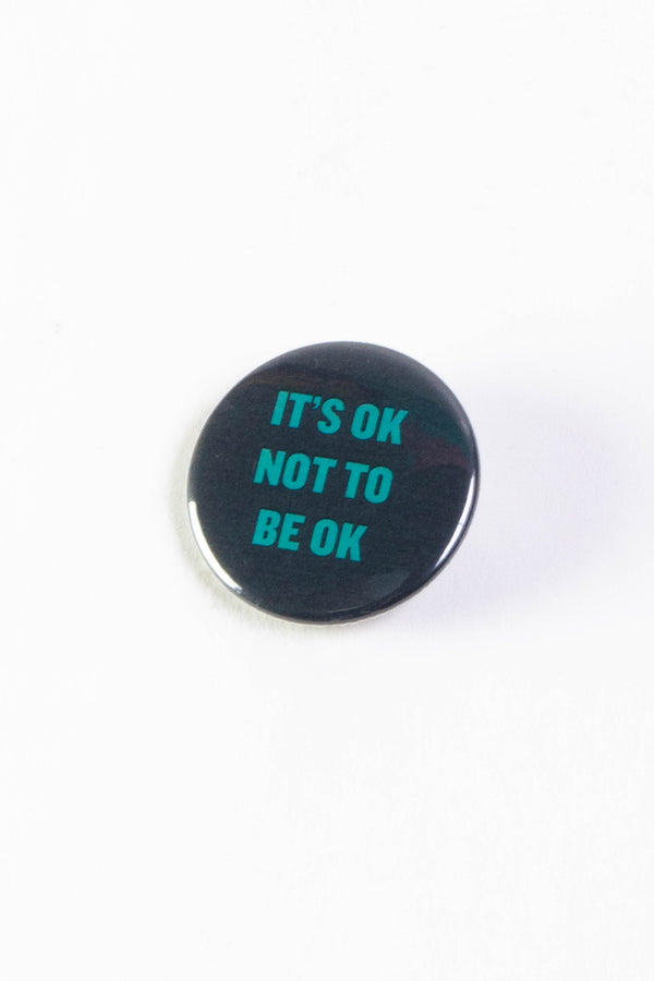 A black pinback button with text that reads "It's ok not to be ok" in teal.