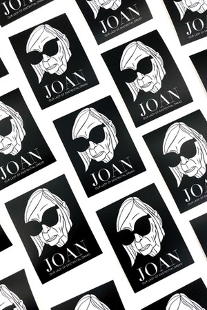 Black rectangle stickers laid out in a tilted grid pattern  with a black and white portrait of Joan Didion's face wearing sunglasses. The sticker says Joan Our Lady of Existential Dread in white text.