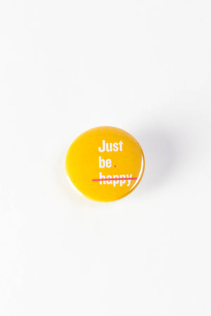 A bright yellow pinback button with white text that reads "Just be happy." A pink line is striking through the word "happy."