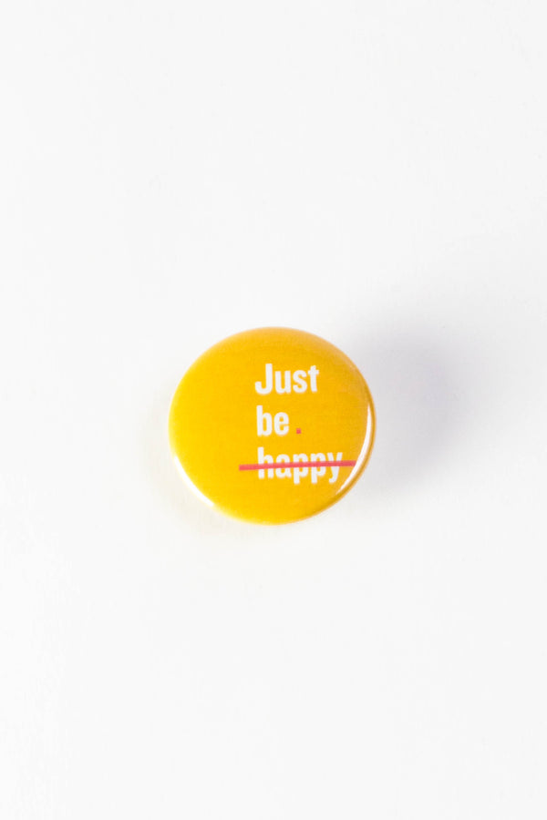 A bright yellow pinback button with white text that reads "Just be happy." A pink line is striking through the word "happy."