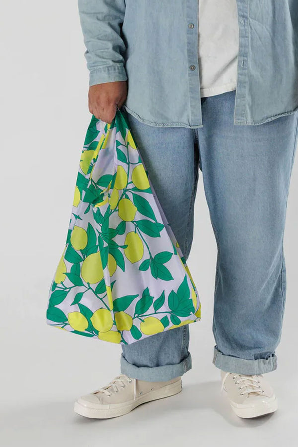 Person holding a white reusable tote bag. The tote bag features lemons and leaves all over the bag. White background.