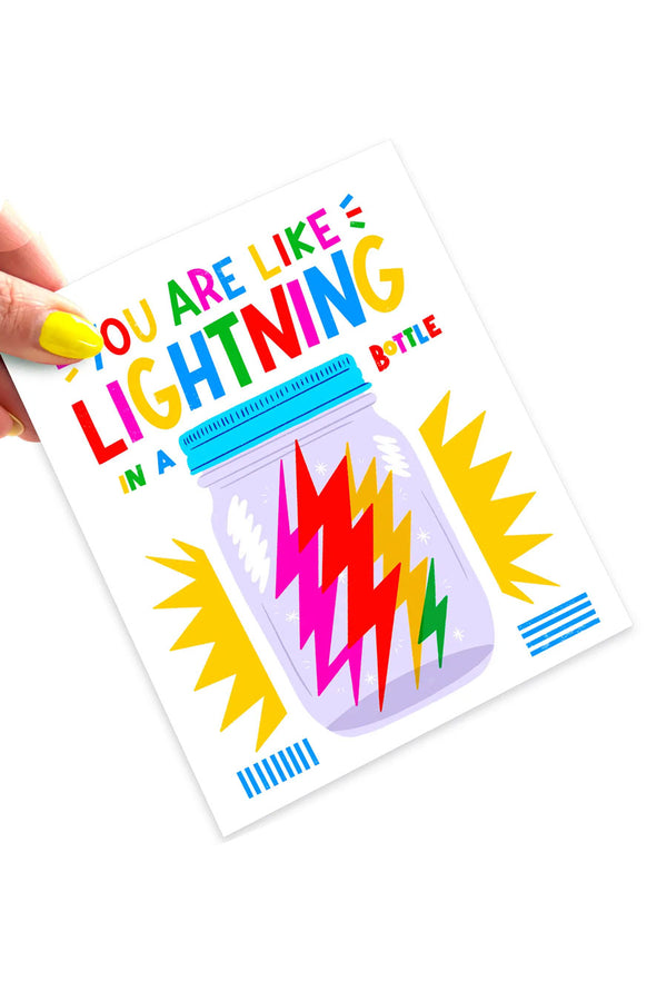 White card against white background. A Person with yellow nail polish holding the card at an angle. The card says "You are like lightning in a bottle" and features an illustration of a mason jar that contains multicolor lightning bolts. Yellow starbusts behind the illustration.