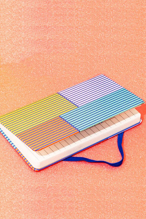 A hard journal with colorblocks of lavender, teal, yellow, and orange features vertical navy stripes laying on its side on a peach glitter background.