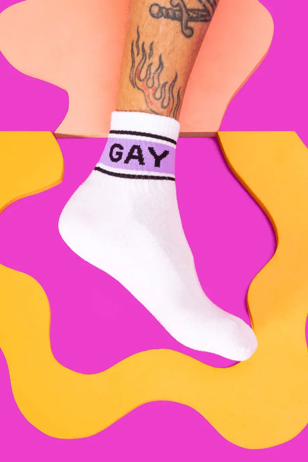 A photo of a persons leg wearing ankle high white crew socks with black stripes and a purple stripe in between. On the purple stripe in black lettering the socks say GAY. Pink background with yellow and peach squiggly lines..