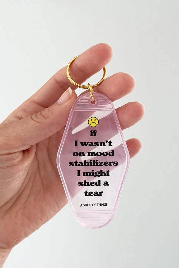 A person holding translucent pink motel style keyring. It says If I wasn't on mood stabilizers I might shed a tear. Above the text is a frown face. Gold keyring at the top. White background.