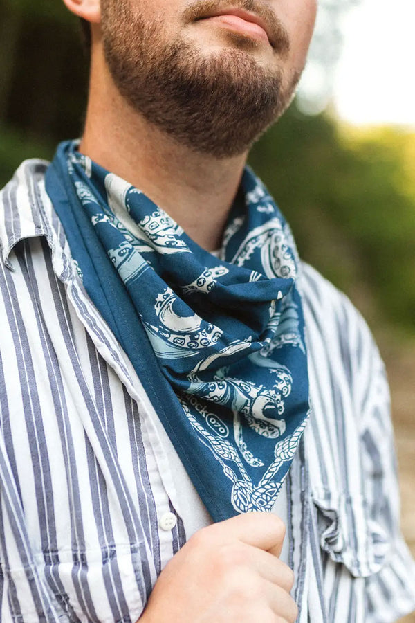 A person wearing a striped button up shirt outside with a navy bandana around their neck. The bandana features octopus tentacles and nautical rope around the border. 
