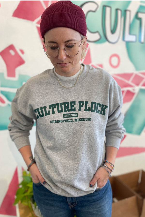 A woman wearing glasses, a burgundy beanie, and a gray sweatshirt with the words "Culture Flock, Est. 2013, Springfield, Missouri" across the chest in green block letters.