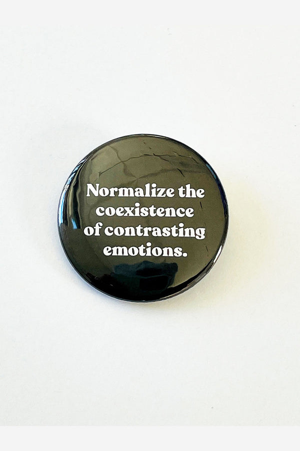 A black pinback button with white text that reads "normalize the coexistence of contrasting emotions."
