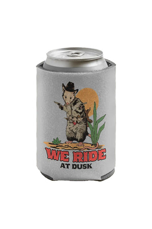 A light gray can koozie sits on a white background. The koozie has an illustration of an opossum dressed as a cowboy holding pistols, standing in the desert. The text reads "We ride at dusk."