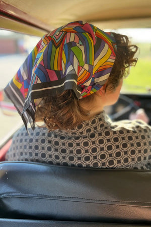 A photo of the back of a persons head with a bandana in their hair. The bandana has a black and white border and features the progress pride, transgender, bisexual, asexual, genderqueer, and nonbinary pride flags.