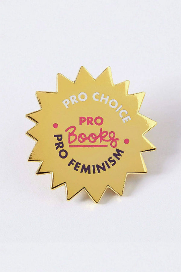 A gold enamel pin in a jagged circle with white, pink, and black text that reads "Pro choice, pro books, pro feminism."