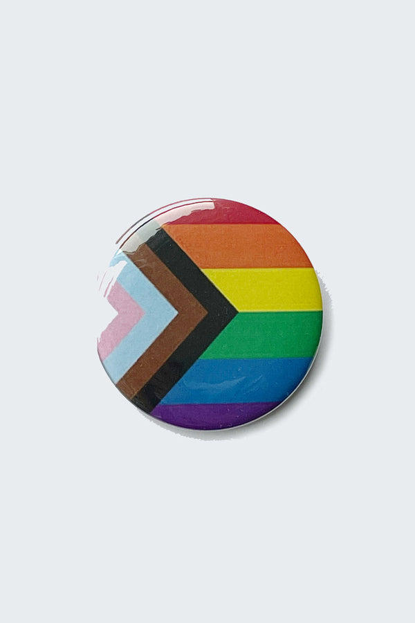 A round pinback button with the image of the Progress pride flag, a modified version of the original pride flag that includes the colors of the transgender flag along with black and brown lines to represent people of color.
