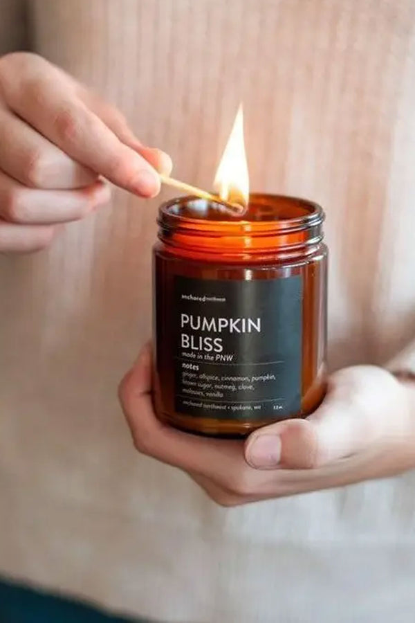 A person holding a candle and lighting it with a match. A brown glass jar candle with a gold lid and a black label that reads "Anchored Northwest, Pumpkin Bliss, made in the PNW. Notes: ginger, allspice, cinnamon, pumpkin, brown sugar, nutmeg, clove, molasses, vanilla.