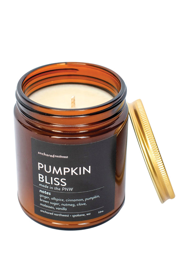 A brown glass jar candle with a gold lid and a black label that reads "Anchored Northwest, Pumpkin Bliss, made in the PNW. Notes: ginger, allspice, cinnamon, pumpkin, brown sugar, nutmeg, clove, molasses, vanilla."