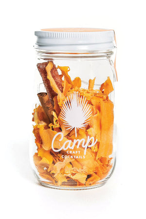 A clear glass mason jar with a white lid is sitting on a flat white surface. The jar is full of dried pumpkin with a cinnamon and a sugar stick. The jar has a white logo that reads "Camp Craft Cocktails."