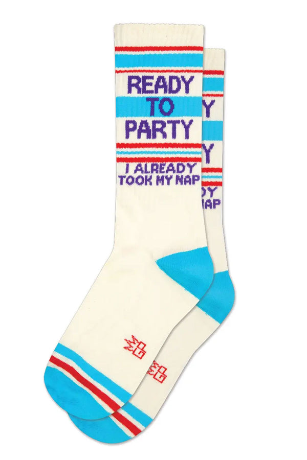Tall mid-calf crew socks. They have a blue heel and toe. There is a blue and red stripe at the toe and around the stop of the socks. In between the stripes at the top of the socks in purple text, it says "Ready to Party I already took my nap". White background.