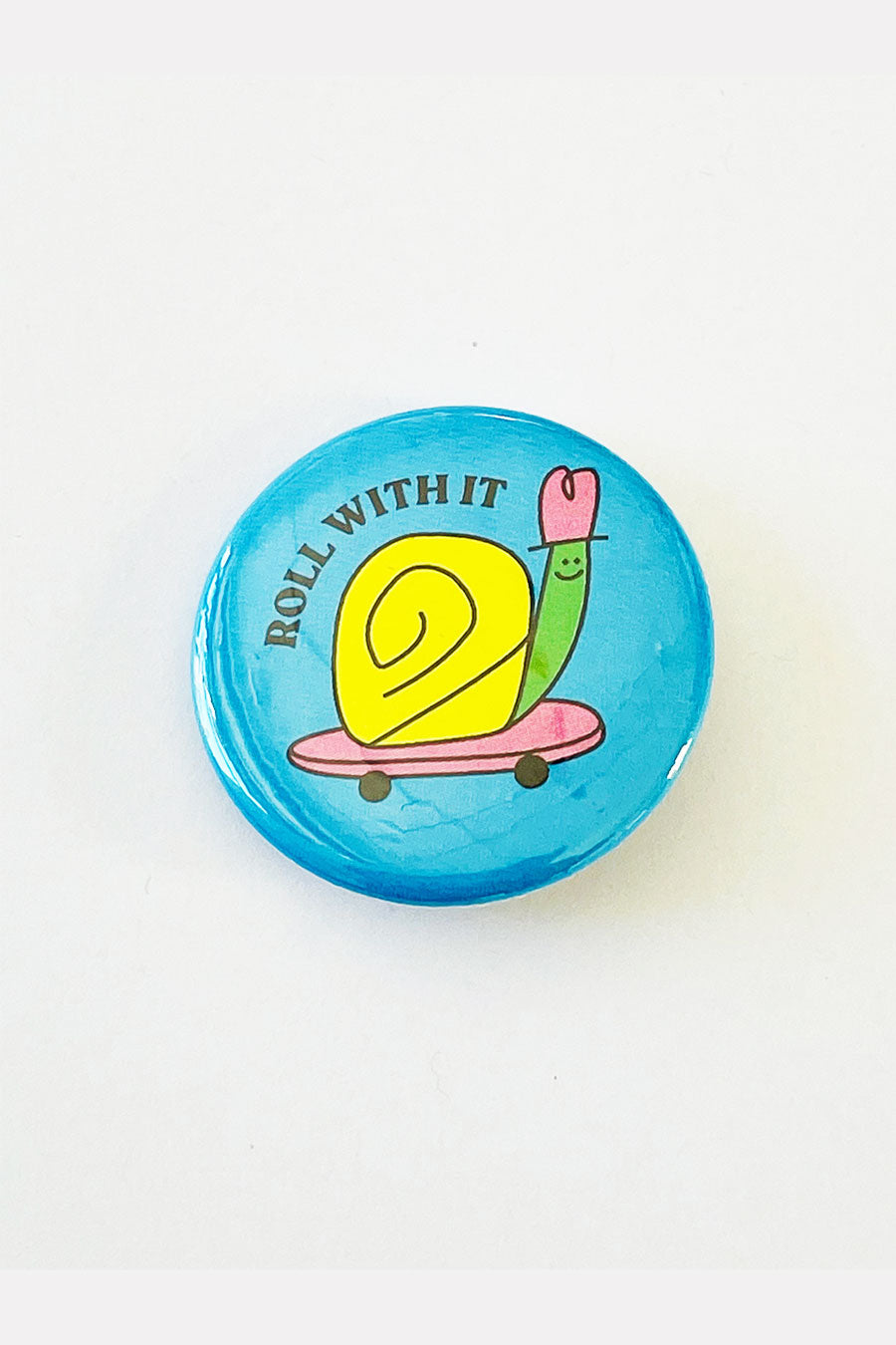 A round light blue pinback button with an illustration of a green and yellow snail riding on a pink skateboard. The black text reads "Roll With It."
