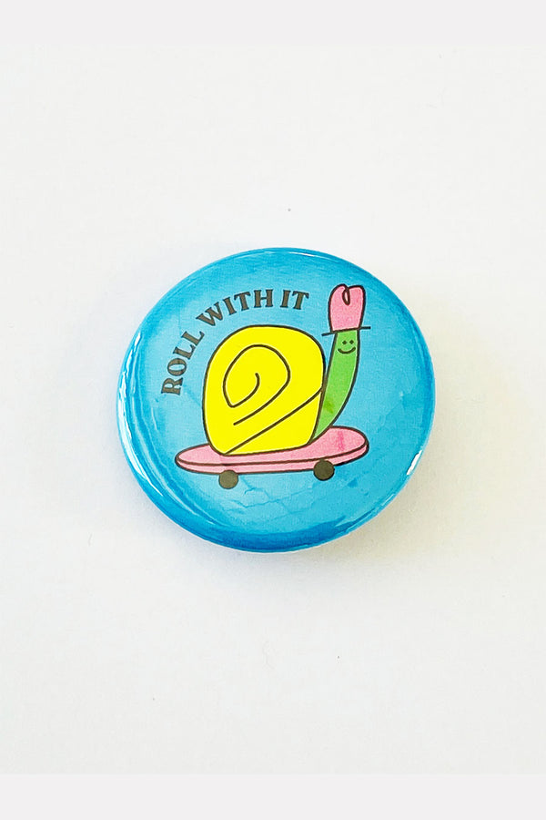 A round light blue pinback button with an illustration of a green and yellow snail riding on a pink skateboard. The black text reads "Roll With It."