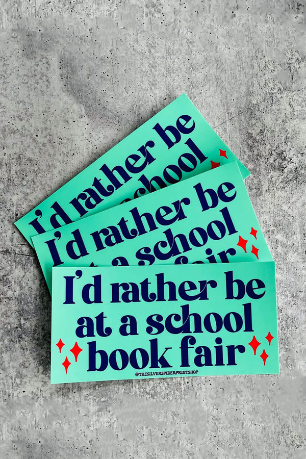 A stack of teal rectangular bumper stickers with navy text that reads "I'd rather be at a school book fair" on a concrete backdrop.