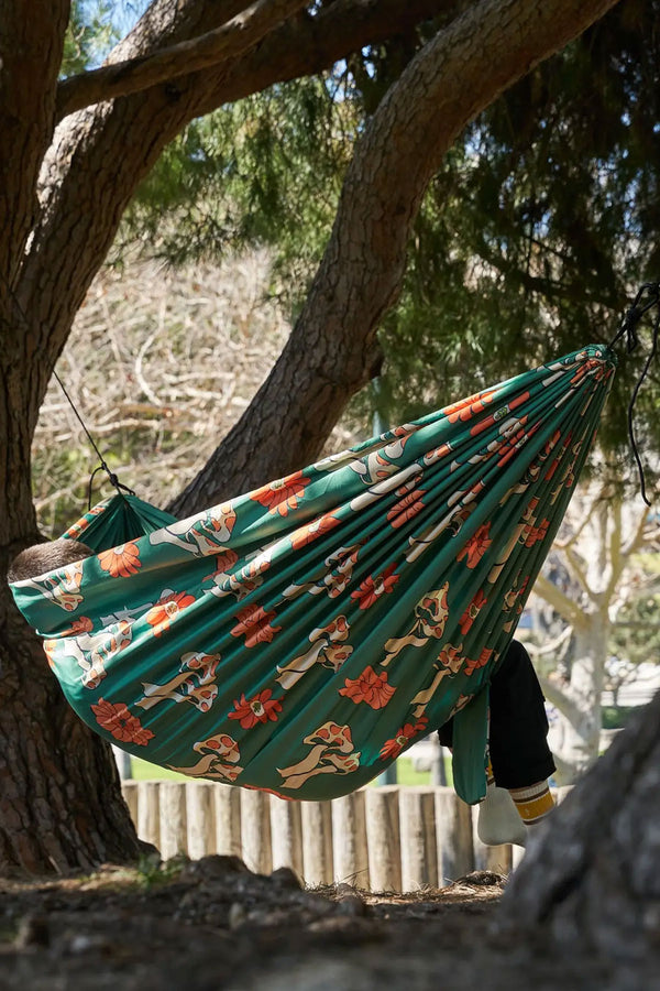 A person sitting under the trees in a two person hammock. The design is a green hammock with white and orange mushroom and flowers printed all over it.