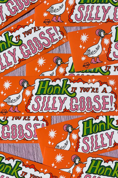 Multiple orange bumper stickers laying on top of one another on a pink linen background. The sticker features a white goose illustration wearing a pink bonnet. The goose has a text bubble coming from its beak that says "Honk if you're a silly goose!" The text is in Green, red, and pink text.