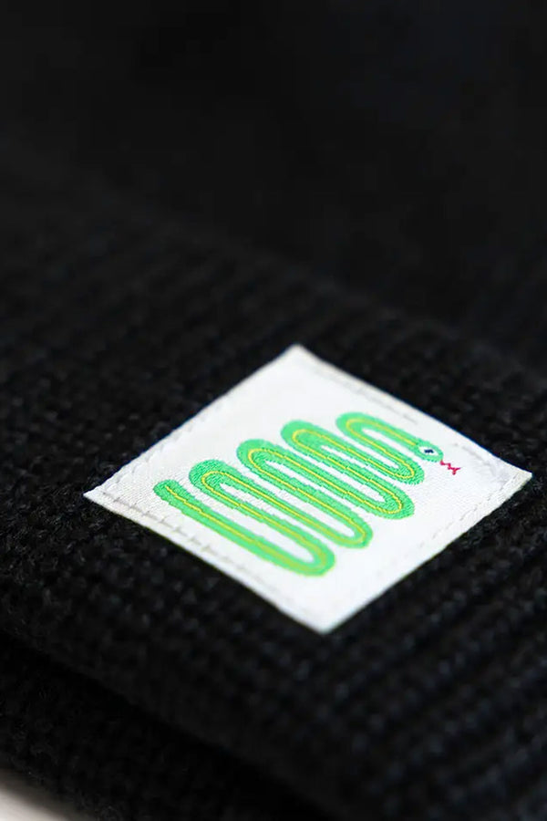 Close up photograph of a square white tag featuring a green snake illustration that is sewn onto a black beanie hat.