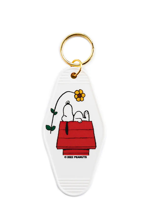 White plastic motel style keychain featuring illustration of snoopy laying on his red doghouse with a daisy leaning over above him. Gold keyring. White background. 