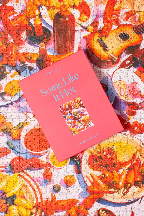 The entire puzzle put together with the puzzle box laid on top. The puzzle features many hot peppers, hot sauces, and a guitar on a light pink tabletop.