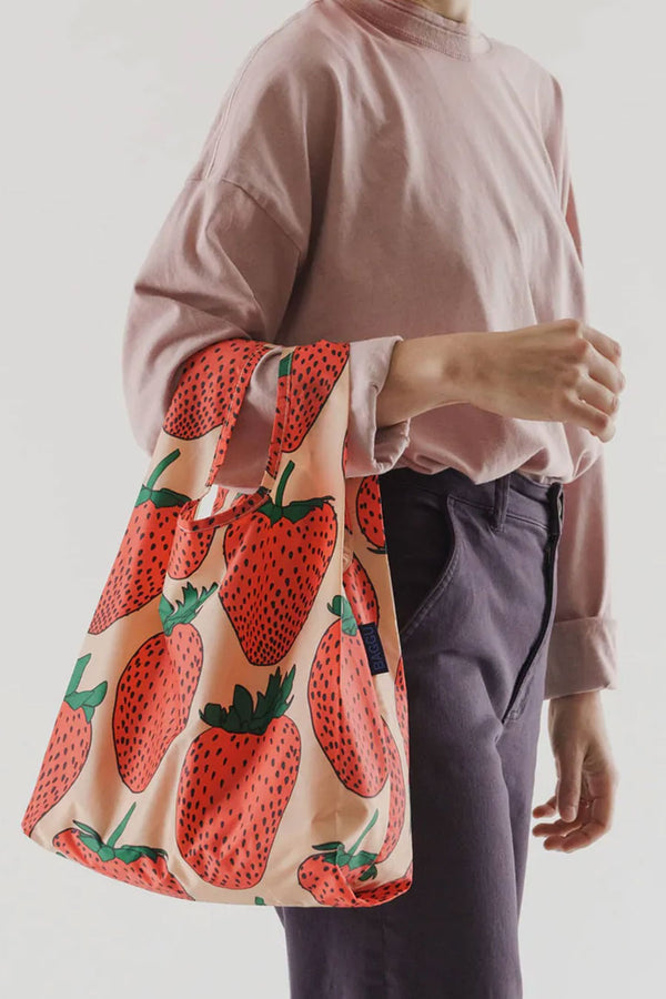 A person standing in front of a white backdrop wearing dark purple pants and a pink long sleeve tee holding a pink reusable tote bag off of their arm. The bag is pink with red strawberries printed all over it.