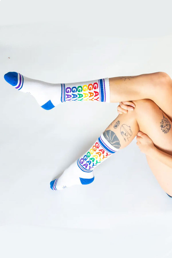 Photo of a persons legs with tattoos wearing tall mid calf white crew socks. The socks have blue heels and toes, blue and purple stripes across the toes and around the top of the socks. The Socks say GAY in repeating pattern in rainbow colors. White background. 