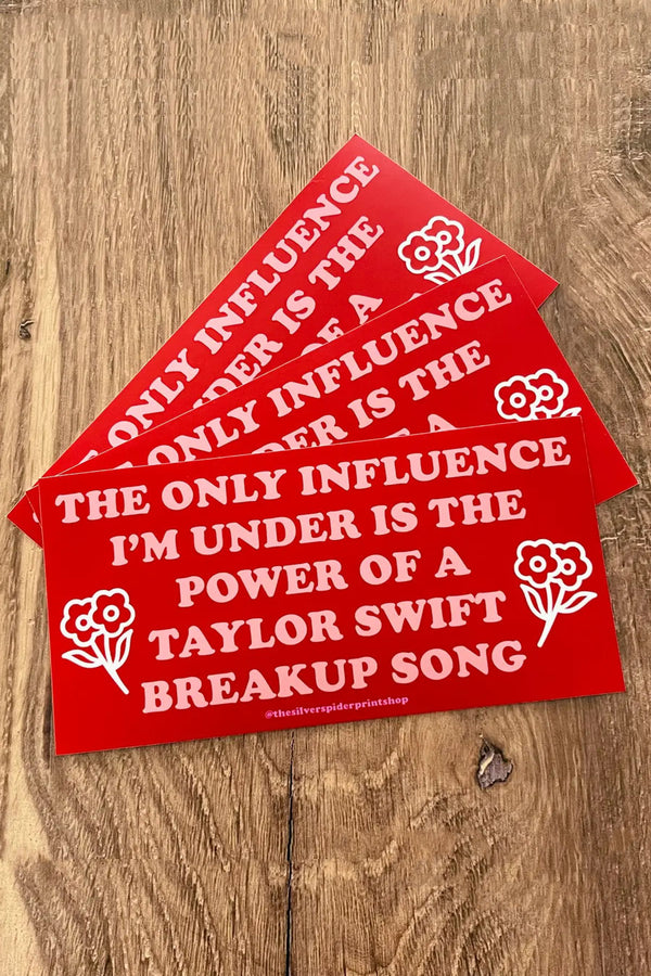 Stack of three red bumper stickers fanned out on a wood surface. The bumper sticker says "The only influence I'm under is the power of a Taylor Swift breakup song" with two small flower clusters on each side of the text.