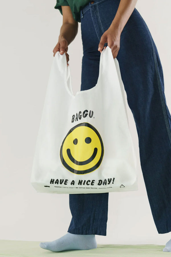 A person in wide leg blue jeans holds white reusable grocery bag from the handles. The bag Says Baggu in black text above a yellow smiley face. Below the smiley face the bag reads "Have a Nice Day!" White background.