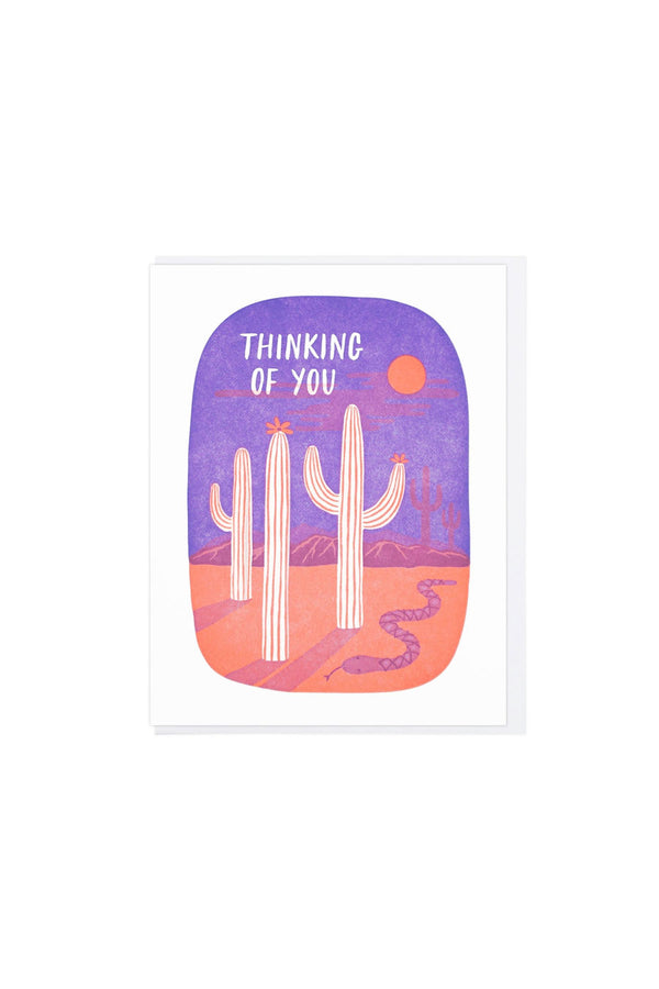 White card on white background. Card features a purple sky and orange desert scene. Three white cacti are in the middle of the card with a sun above them. A small snake to the right of the cacti. The card says "Thinking of you" in white lettering against the purple sky.